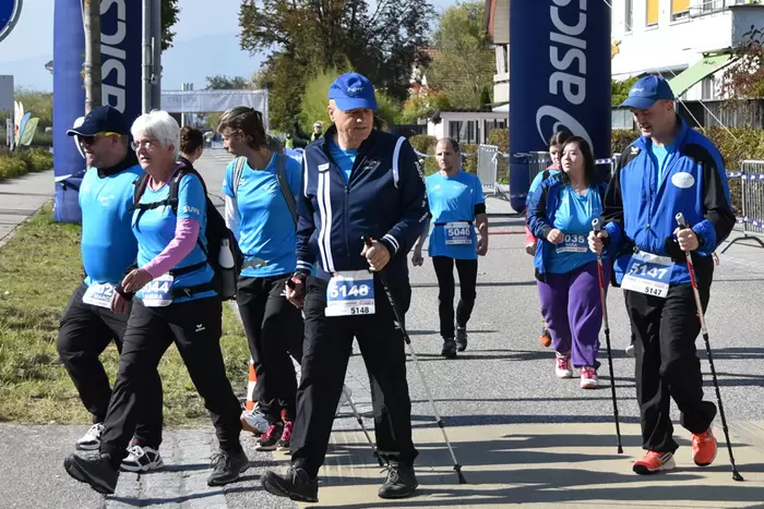 Eine Gruppe von Menschen in Sportkleidung nimmt an einem Wander- oder Nordic-Walking-Event im Freien teil. Sie tragen nummerierte Startnummern und benutzen Wanderstöcke an einem sonnigen Tag.